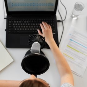 Eine Frau sitzt vor einem Mikrofon und Laptop und bearbeitet einen eingesprochenen Text. Daneben liegt auf dem Tisch liegen Blätter mit markiertem Text und darüber steht ein Wasserglas.