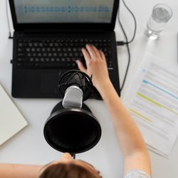Eine Frau sitzt vor einem Mikrofon und Laptop und bearbeitet einen eingesprochenen Text. Daneben liegt auf dem Tisch liegen Blätter mit markiertem Text und darüber steht ein Wasserglas.